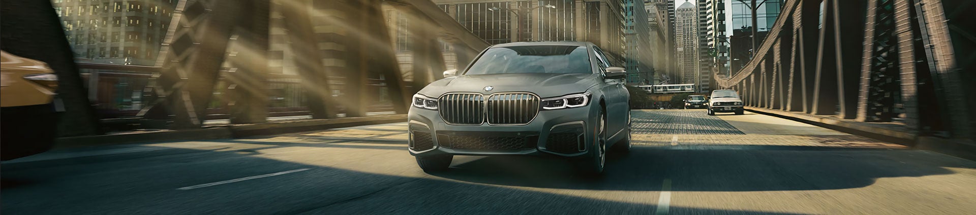 A Bernina Gray Amber Effect BMW 7 Series driving across city bridge.